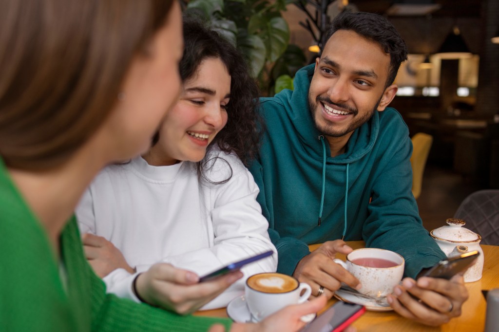 Three young adults enjoying drinks and using their phones at a café table, smiling and engaged in conversation.